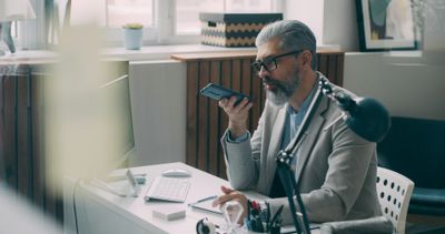 a person sitting at a desk with a computer, phone in hand, as if to talk to an AI recording tool on his phone.