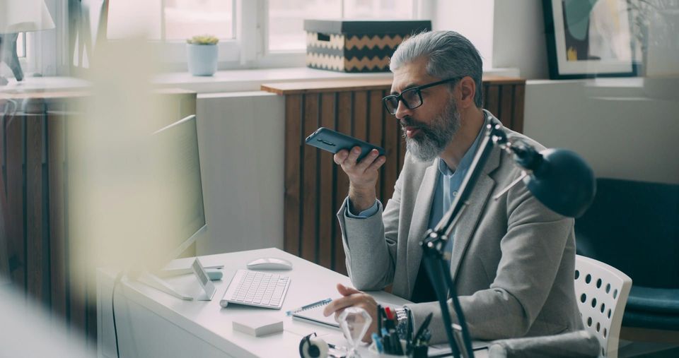 a person sitting at a desk with a computer, phone in hand, as if to talk to an AI recording tool on his phone.