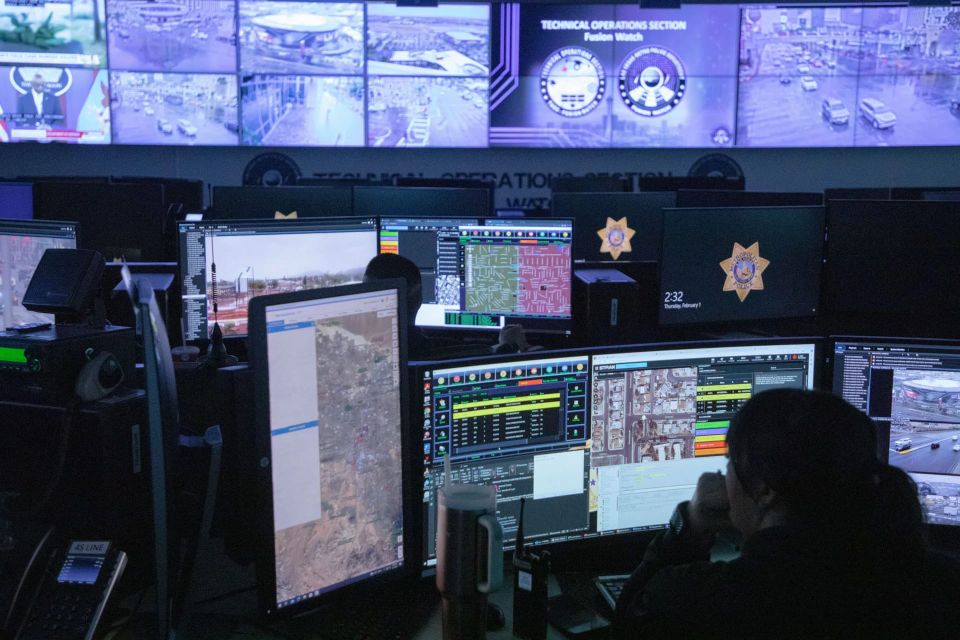 a photo of a dark room as a fusion technical operations center, with tiled displays on the far wall and several agents sitting at computers with blurry intelligence and maps on their screens.