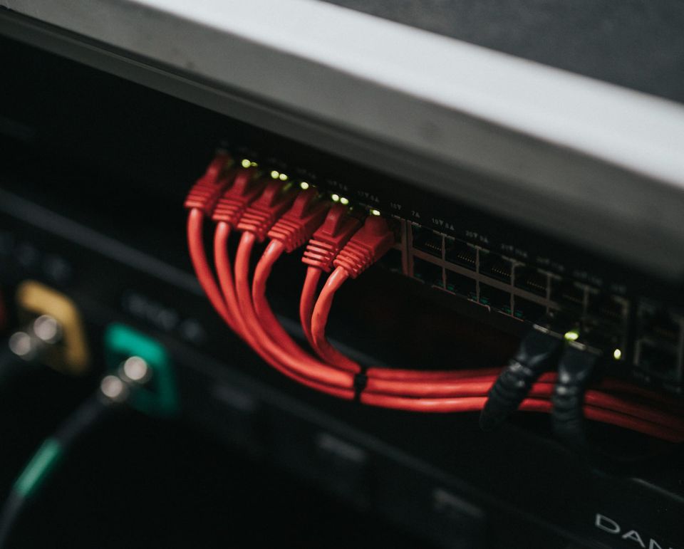a photo of a bunch of red ethernet cables plugged into ports on a router, in a server rack in a darkened datacenter room; the lights are flashing on the back of the ethernet cables.