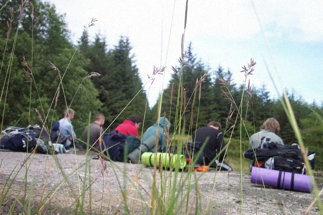 Six lads resting during a long hike. They are sitting on a bridge, their packs behind them. The foreground is tall grass.