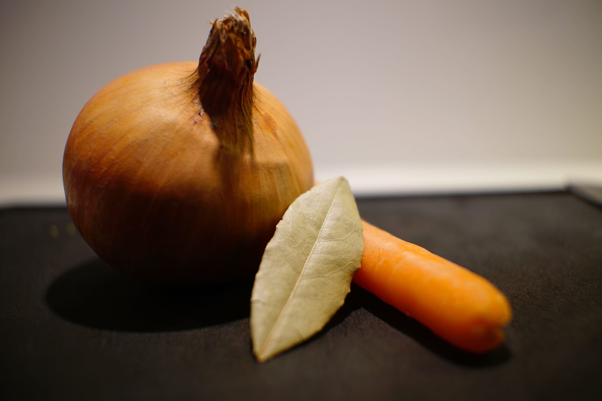 An onion, a carrot, and a bay leaf on a chopping board.