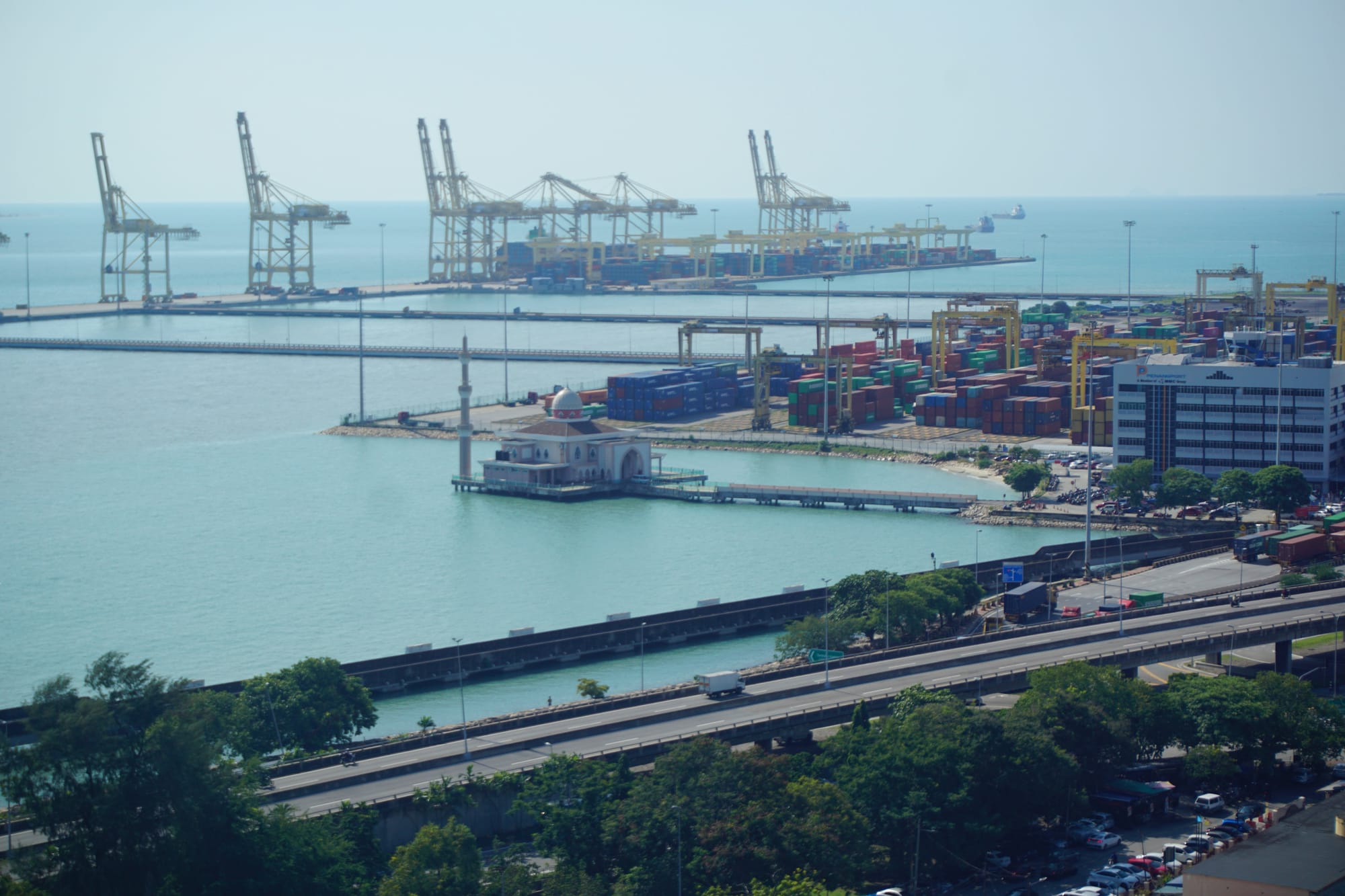 A view over Penang port, with cranes loading a container ship in the back, many containers stacked in the mid, and the mosque on a platform to the side.