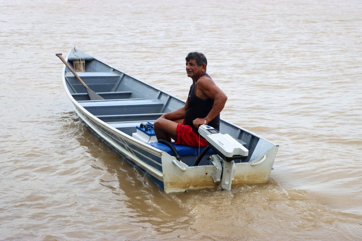 Pescador em uma canoa de alumínio com um motor de popa elétrico branco, navegando em um rio de águas barrentas.