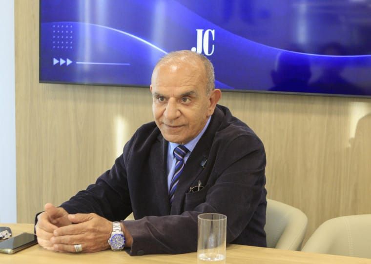 O candidato Milton Cardoso, de terno escuro e gravata listrada, gesticula durante entrevista em frente a uma tela azul.