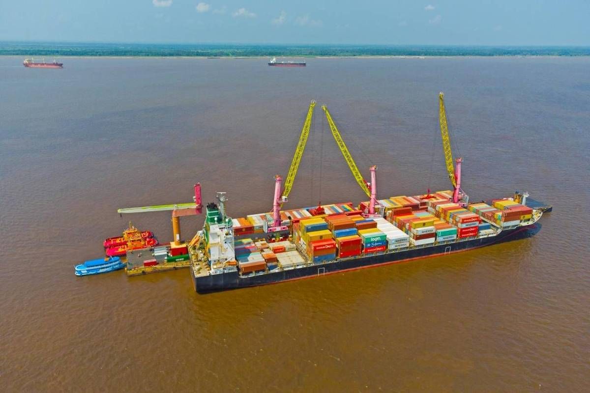 Navio cargueiro com contêineres coloridos sendo descarregados por guindastes em um píer flutuante no rio, com outras