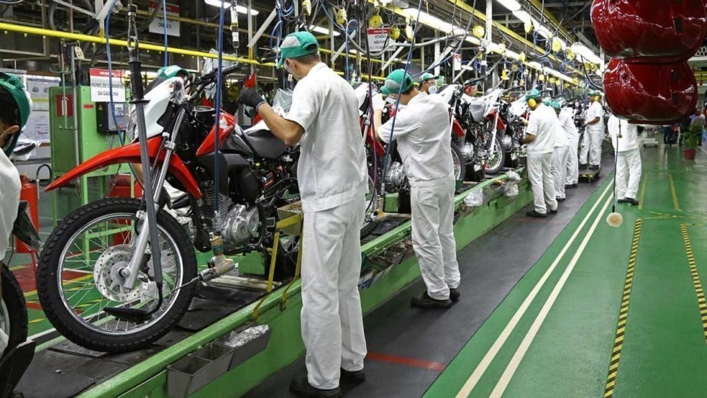 Trabalhadores com uniformes brancos e bonés verdes montam motocicletas vermelhas em linha de produção da Honda em Manaus.