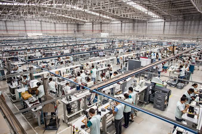 Vista aérea de uma fábrica com dezenas de trabalhadores em suas estações de trabalho, realizando tarefas de montagem