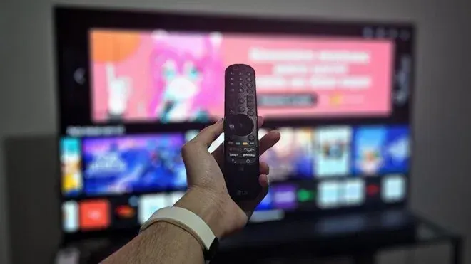 Mão segurando controle remoto da LG em frente a uma Smart TV ligada exibindo menu de aplicativos e canais.