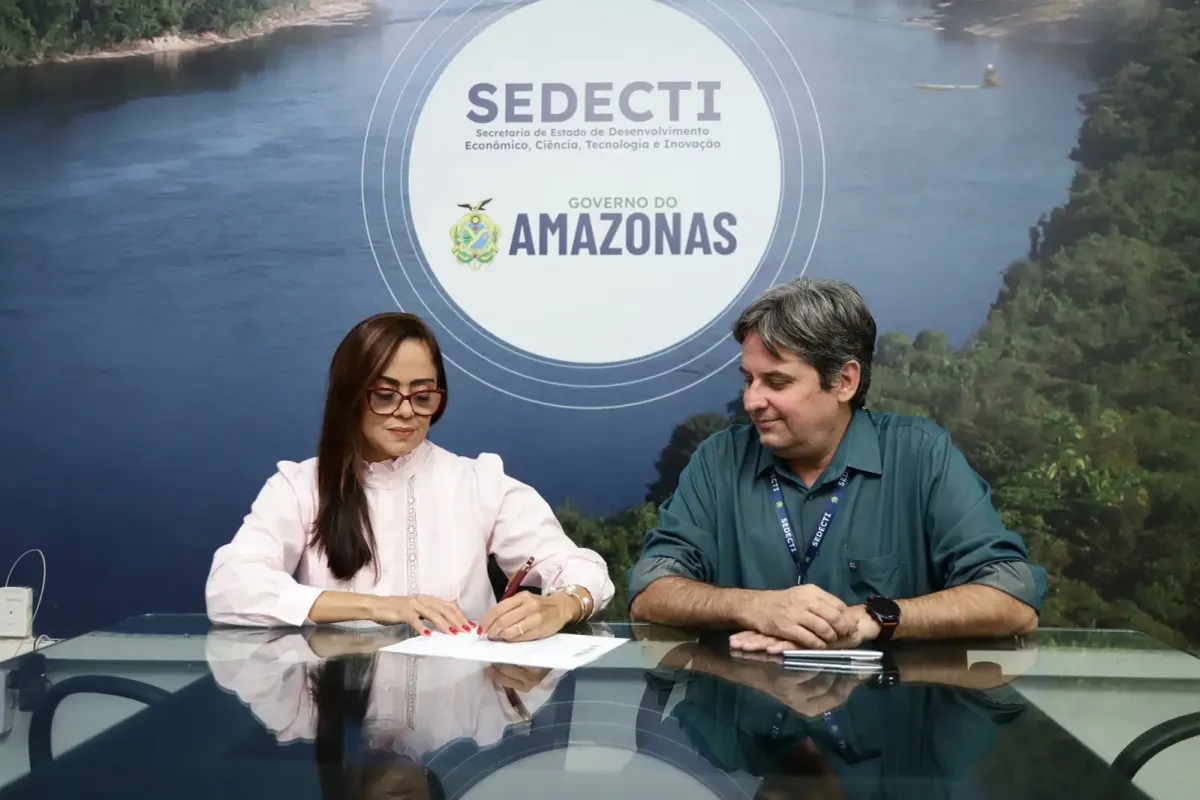 Luzia Raquel Said assina documento ao lado de um homem, com o logo da Sedecti e do Governo do Amazonas ao fundo, em Manaus