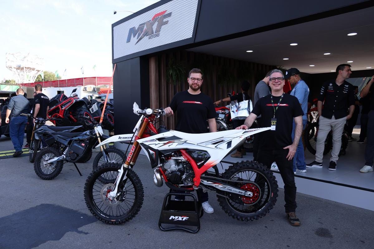 Dois homens sorrindo ao lado de uma motocicleta MXF branca e vermelha em um estande da MXF Motors