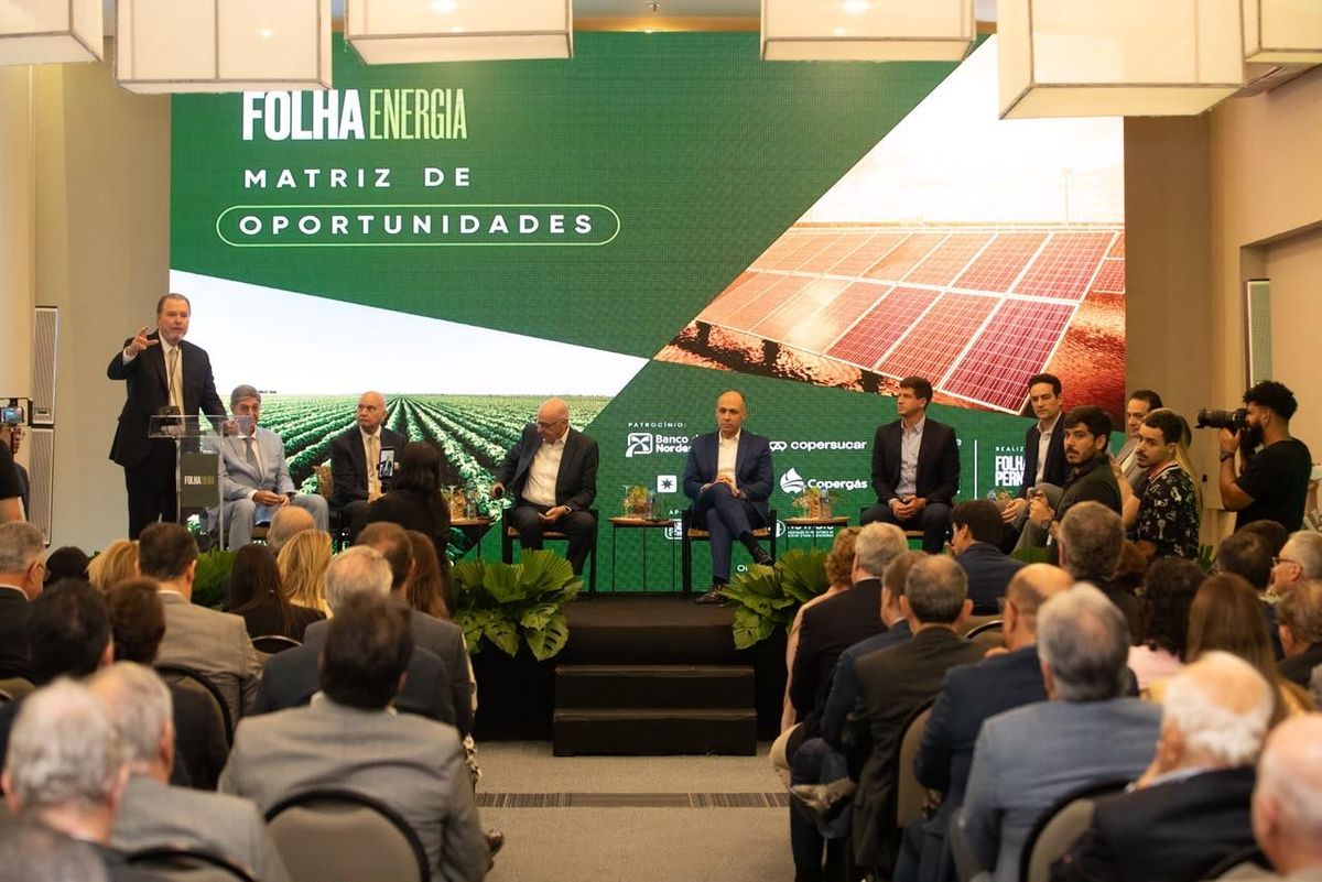 Homem discursa em púlpito durante o evento Folha Energia, com painel ao fundo exibindo plantação e placas solares.