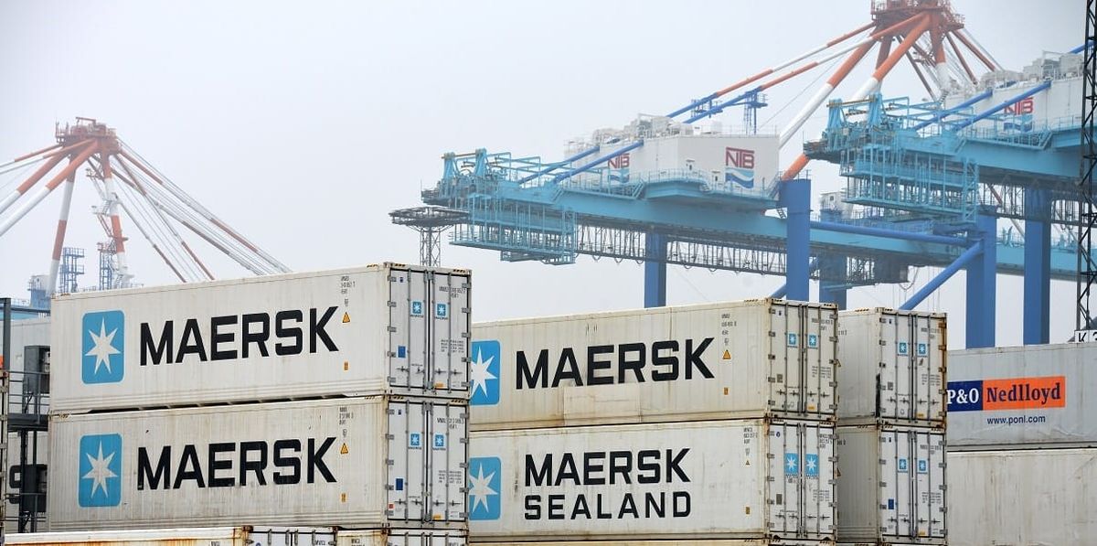 Contêineres brancos da Maersk empilhados em um terminal portuário com guindastes azuis ao fundo.