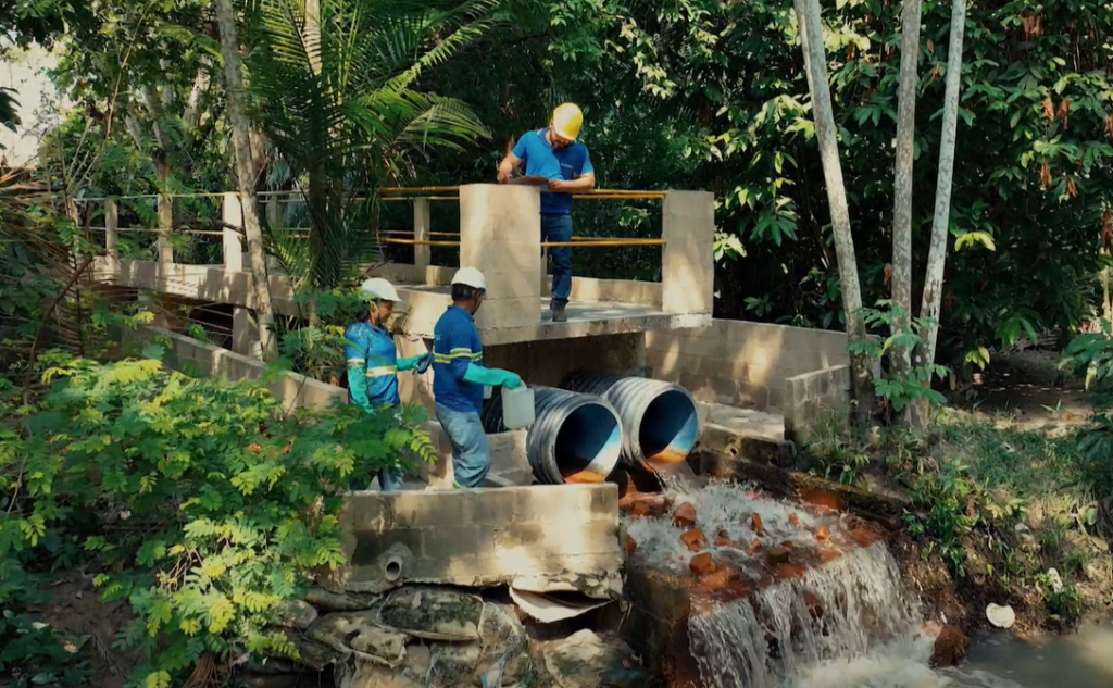 Três técnicos com capacetes e uniformes azuis realizam coleta de amostras em tubulações de esgoto em área de mata em Manaus.