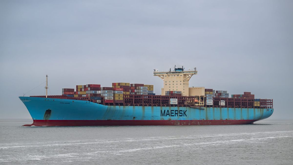Navio de carga da Maersk, azul com casco vermelho, carregado de contêineres coloridos, navegando em águas calmas sob céu nublado