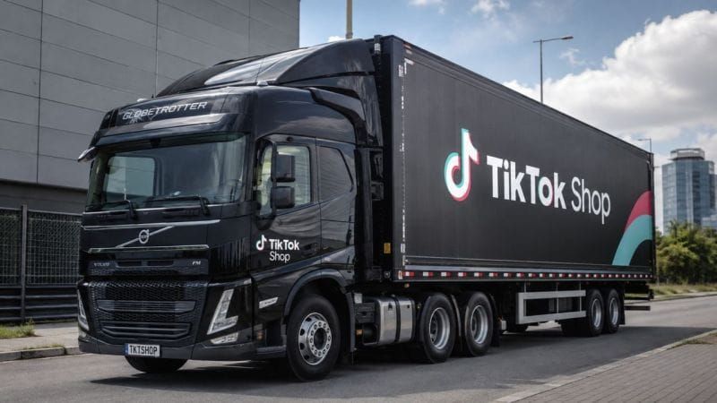 Caminhão preto da Volvo com a marca TikTok Shop na lateral do baú e na cabine, estacionado em frente a um prédio cinza