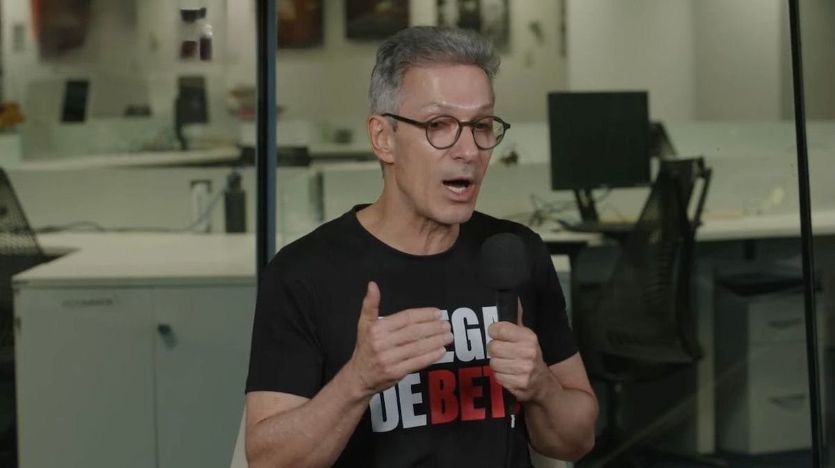 Romeu Zema fala ao microfone durante entrevista em um escritório, vestindo camiseta preta com inscrições em branco e vermelho.