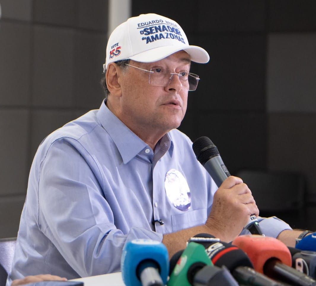Senador Eduardo Braga fala ao microfone durante entrevista coletiva, usando boné branco e camisa azul.