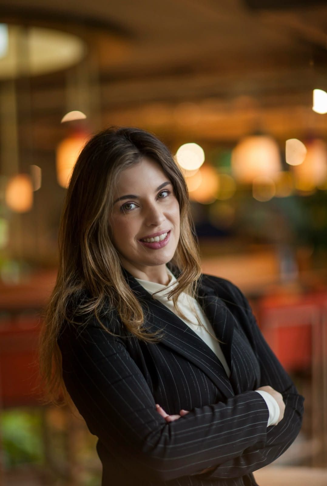 A economista Karine Atala Frota sorrindo, vestindo um blazer preto risca de giz, em um ambiente interno iluminado.