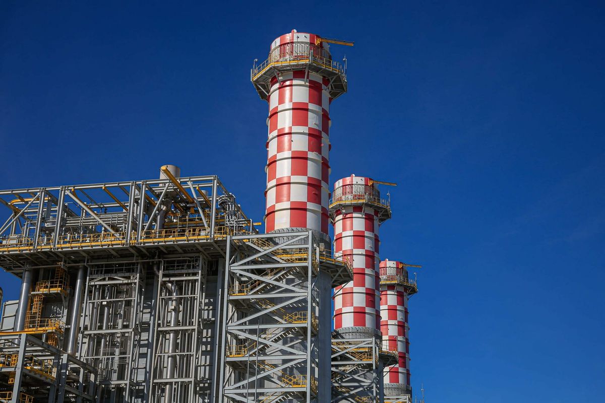 Três chaminés industriais listradas em vermelho e branco sob céu azul em uma usina termelétrica.