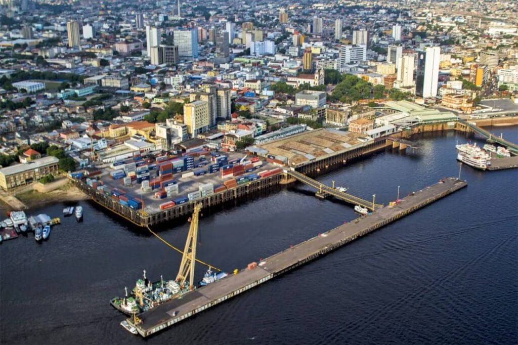 Vista aérea do Porto de Manaus com contêineres coloridos, píer e embarcações no Rio Negro, tendo a cidade ao fundo.