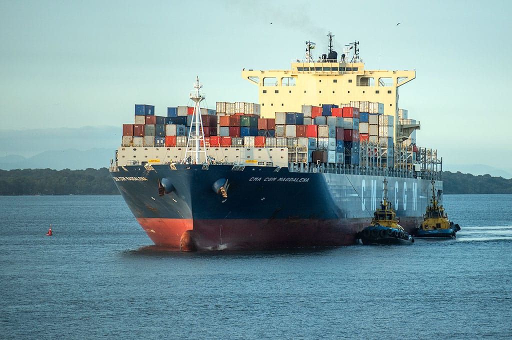 Navio porta-contêineres da CMA CGM navegando em águas calmas, acompanhado por dois rebocadores laterais.