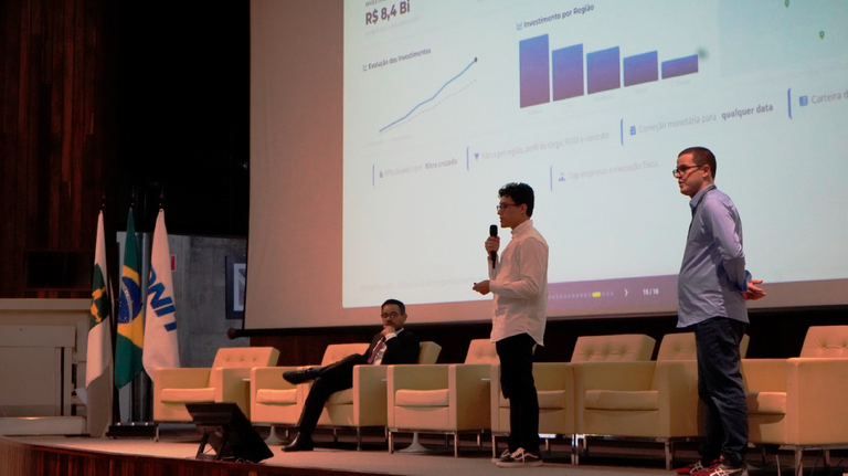 Homem apresenta novo sistema da Antaq em palco com projeção de gráficos de investimentos e bandeiras ao fundo.