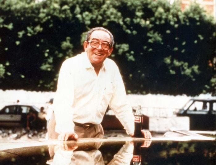 O empresário Antônio Simões sorrindo, usando óculos e camisa clara, apoiado em uma superfície com uma garrafa de Coca-Cola.