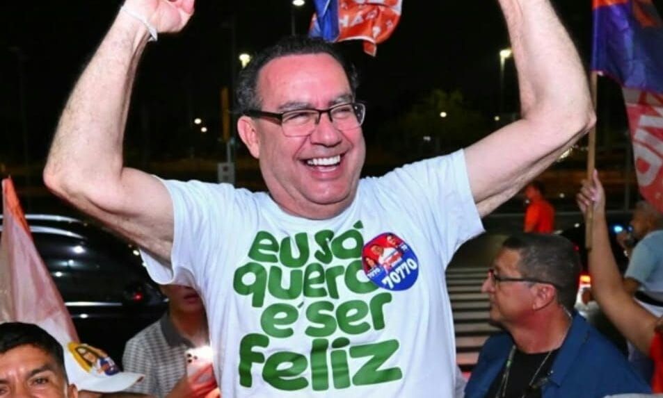 Augusto Cury sorrindo com braços erguidos, vestindo camiseta branca com a frase eu só quero é ser feliz em letras verdes.