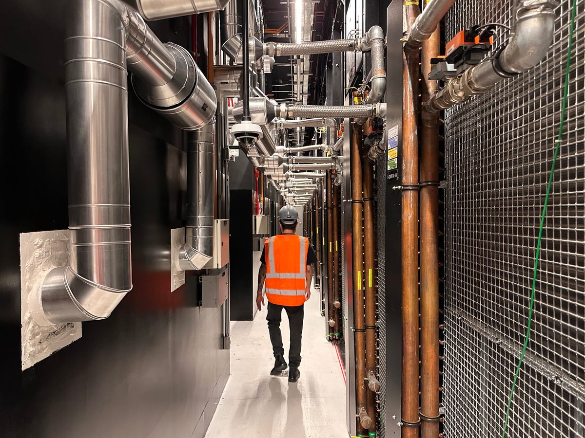 Trabalhador com colete reflexivo caminha por corredor técnico de datacenter com tubulações industriais e cabos expostos.
