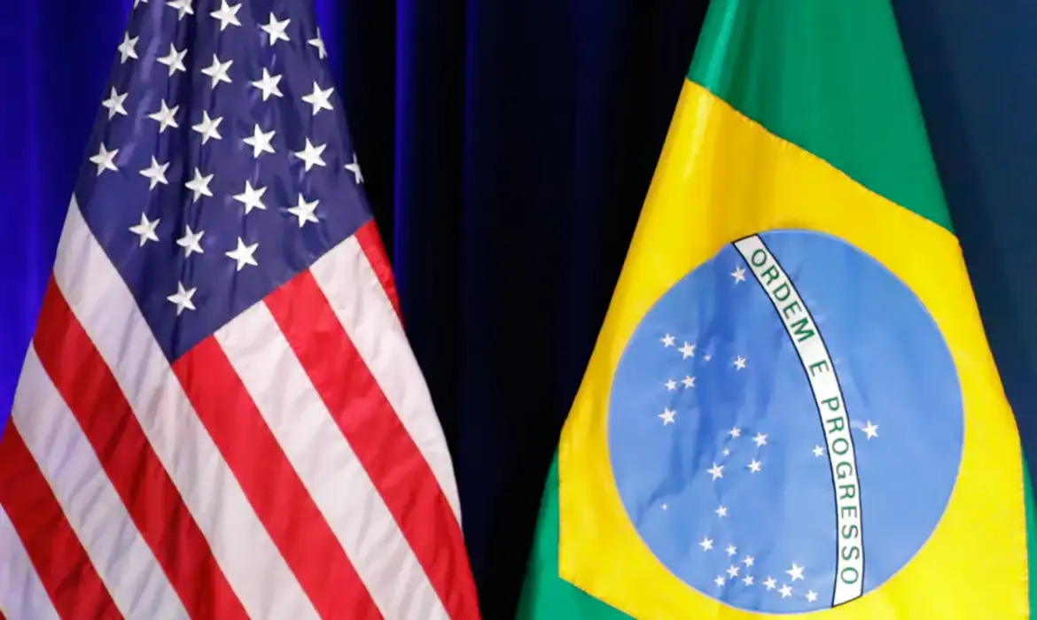 Bandeiras dos Estados Unidos e do Brasil posicionadas lado a lado em um ambiente interno.