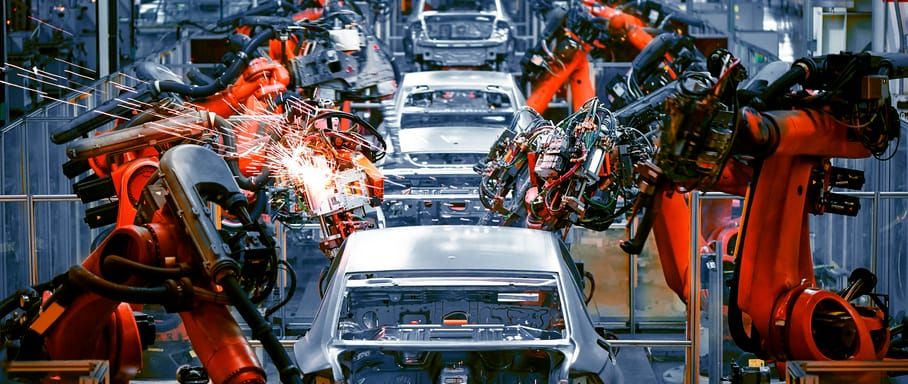 Braços robóticos industriais de cor laranja realizam soldagem em carcaça de veículo em linha de montagem automatizada.