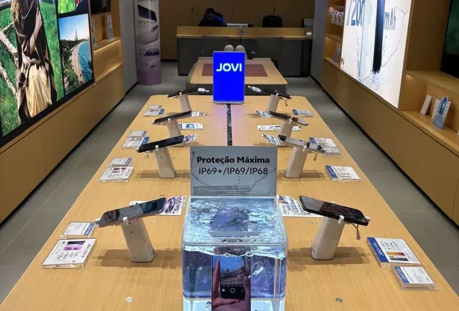 Smartphones da marca Jovi expostos em bancada de loja, com destaque para teste de resistência à água em recipiente de vidro.