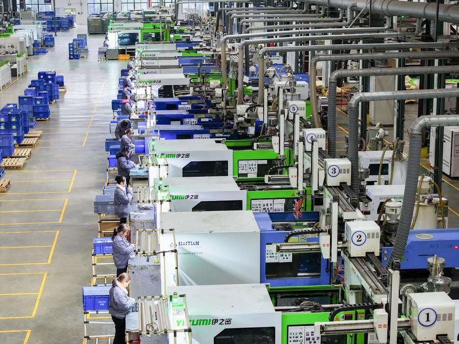 Linha de produção industrial com máquinas automatizadas e operários em uniformes cinza operando equipamentos numerados.