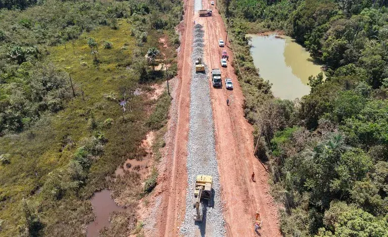 Vista aérea de máquinas e caminhões trabalhando em trecho de terra da rodovia BR-319 cercada por vegetação e água.