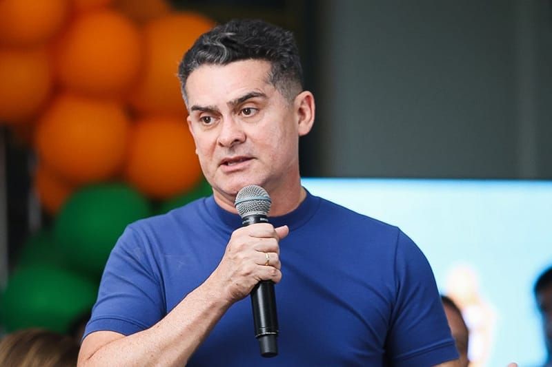 David Almeida fala ao microfone durante evento, vestindo camiseta azul, com balões laranjas e verdes ao fundo.