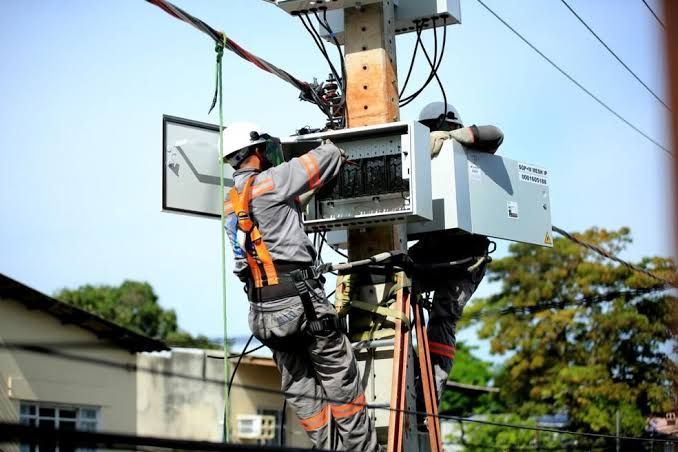 Dois eletricistas com equipamentos de segurança trabalham na manutenção de medidores de energia em um poste em Manaus.