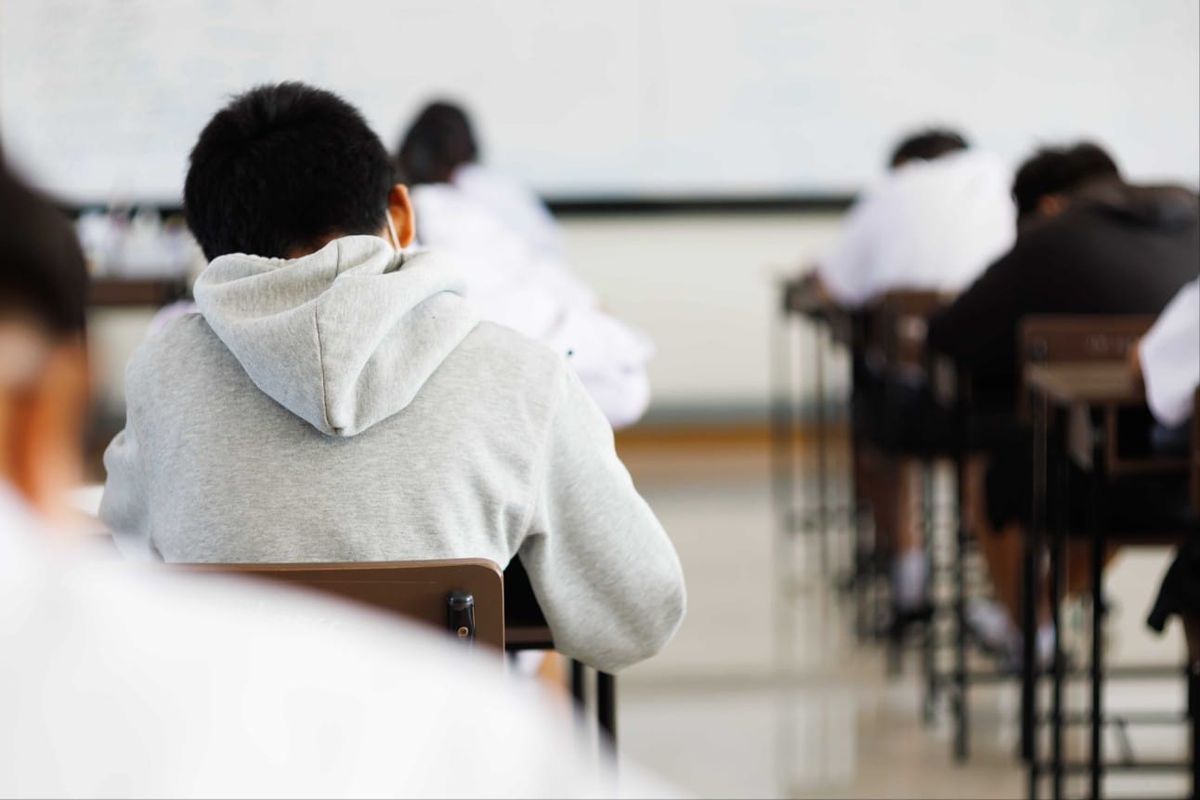 Estudantes sentados em carteiras escolares dentro de uma sala de aula, vistos de costas.
