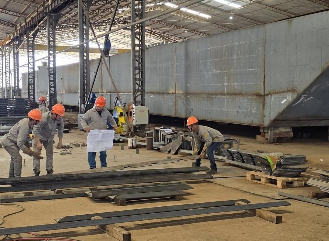 Trabalhadores com capacetes e uniformes manuseiam peças de metal dentro de um galpão industrial no Estaleiro Juruá.