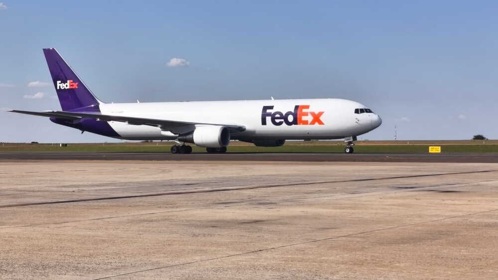 Avião de carga branco com a logomarca roxa e laranja da FedEx estacionado na pista de um aeroporto sob céu azul.