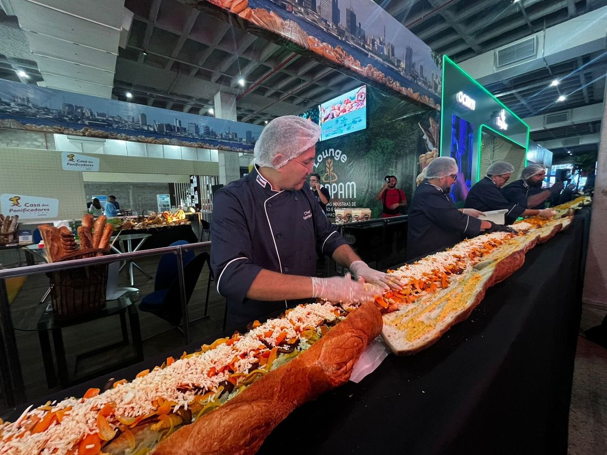 Homens com uniformes de chef e toucas montando um sanduíche gigante em uma mesa comprida durante evento de panificação.