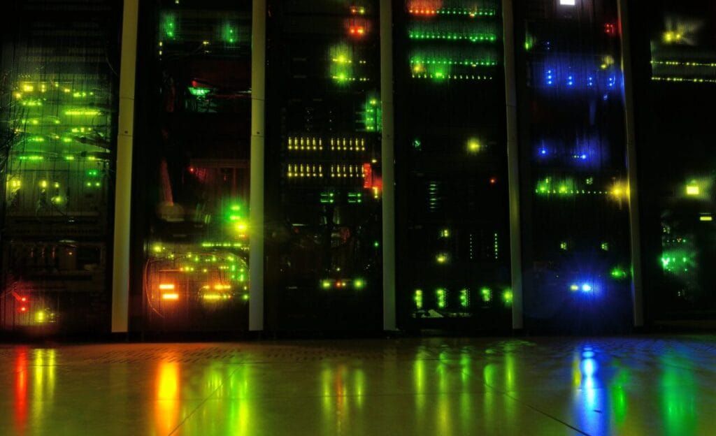 Servidores de um data center com luzes verdes, amarelas e azuis refletidas no chão de uma sala escura.