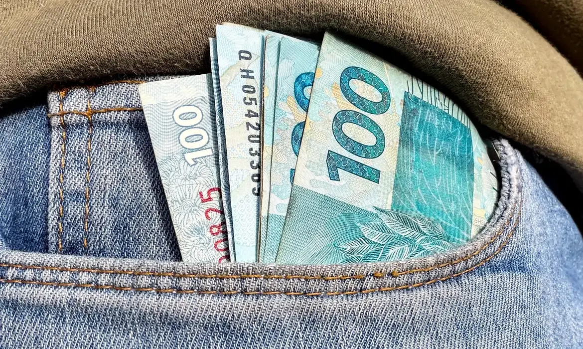 Cédulas de cem reais dobradas dentro do bolso de uma calça jeans azul.