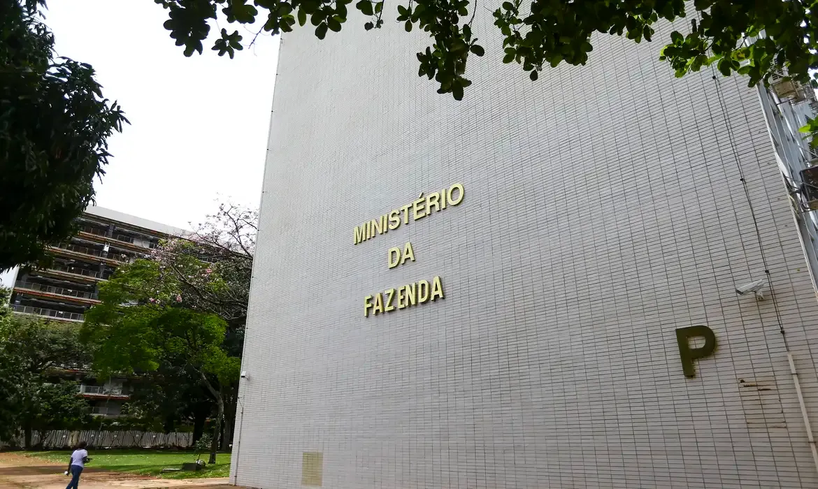 Fachada do edifício do Ministério da Fazenda com letreiro dourado em parede branca e árvores ao redor.