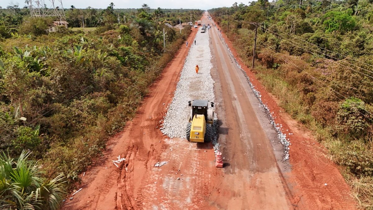 Rolo compressor amarelo trabalha em trecho de terra e brita da rodovia BR-319 cercada por vegetação amazônica.
