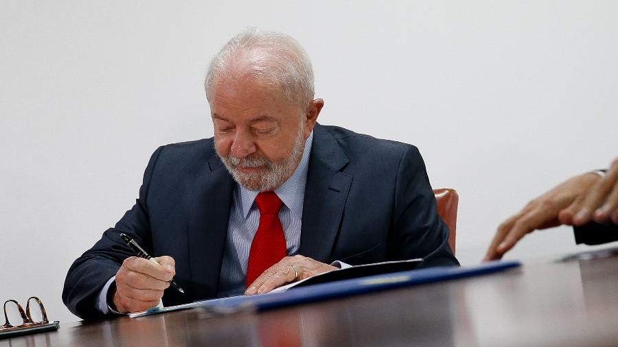 O presidente Luiz Inácio Lula da Silva assina documento oficial sobre uma mesa de madeira.