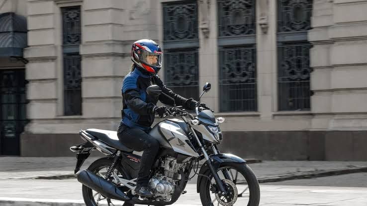 Pessoa pilotando uma motocicleta Honda prateada em uma via urbana em frente a um prédio histórico.
