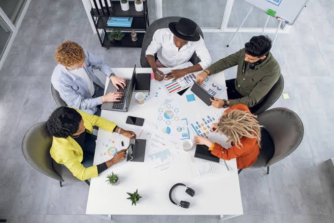 Cinco pessoas sentadas ao redor de uma mesa branca em reunião, analisando gráficos e usando notebooks e tablets.