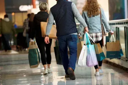 Pessoas caminhando em um centro comercial carregando sacolas de compras de diversos tamanhos e cores.
