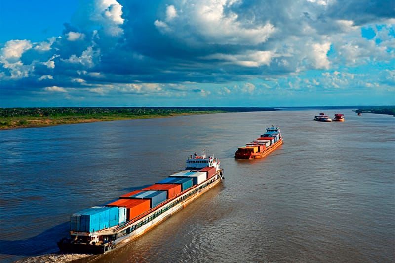 Balsas carregadas com contêineres coloridos navegando pelo Rio Amazonas sob céu com nuvens carregadas.